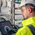man, an electrical technician working in a switchboard with fuses, uses a tablet.